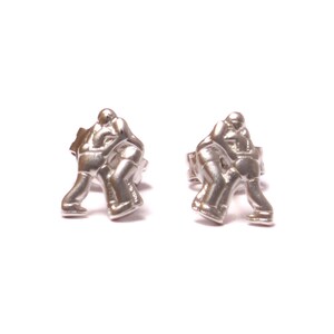 May include: A pair of silver stud earrings featuring a design of two figures wrestling. The earrings are a simple and stylish accessory.