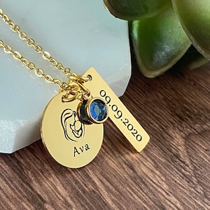 Yellow Gold Necklace engraved with an image of a mom holding a baby and personalized with the daughter's name Ava. Next is a rectangle tag with the birthdate 9.9.20 and a blue September birthstone. Charms are hung on a gold cable chain.