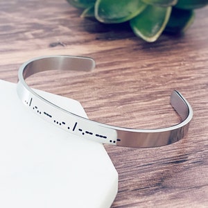 May include: A silver bangle bracelet with a message in braille. The bracelet is on a white surface.