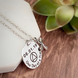 May include: Silver necklace with a round pendant that says "06.01.19 One Day at a Time" with a triangle symbol and a small silver cross charm.