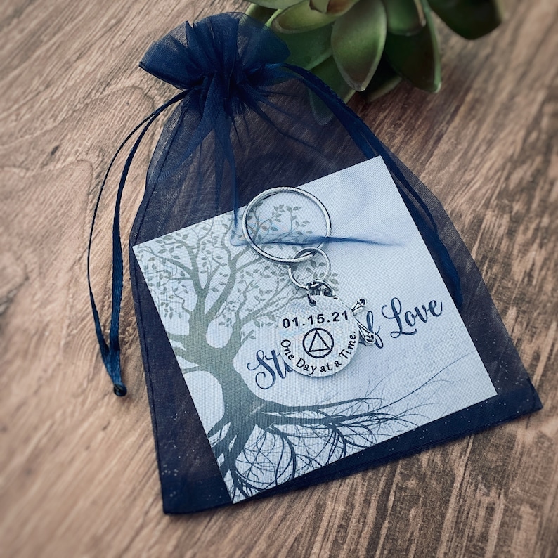 May include: A silver keychain with a round charm that says "One Day at a Time" and "01.15.21" with a triangle symbol. The keychain is in a blue organza bag with a card that says "Strength of Love" and has a tree design.