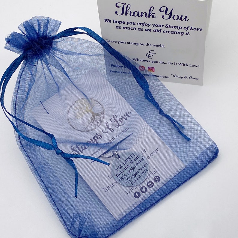 K&ouml;nnte beinhalten: Eine blaue Organzabtasche mit einem silbernen Anh&auml;nger. Der Anh&auml;nger ist rund und tr&auml;gt den Text "I'M LOST! Call my Mom!" Die Tasche liegt auf einer wei&szlig;en Karte mit dem Text "Stamps of Love" und einer Website-Adresse.