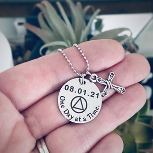 May include: Silver necklace with a round pendant that says "08.01.21 One Day at a Time" with a triangle symbol in the center. A small silver cross charm hangs from the pendant.