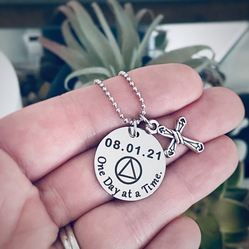 May include: Silver necklace with a round pendant that says "08.01.21 One Day at a Time" with a triangle symbol in the center. A small silver cross charm hangs from the pendant.