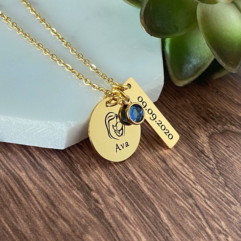 Yellow Gold Necklace engraved with an image of a mom holding a baby and personalized with the daughter's name Ava. Next is a rectangle tag with the birthdate 9.9.20 and a blue September birthstone. Charms are hung on a gold cable chain.