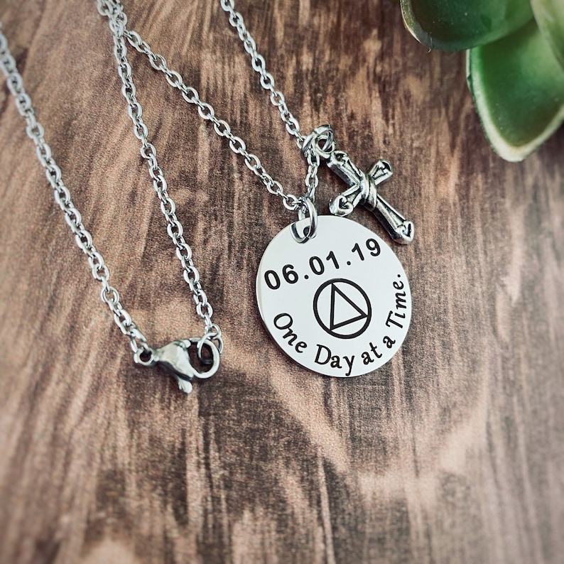 May include: Silver necklace with a round pendant that says "06.01.19 One Day at a Time." The pendant also features a triangle symbol. The necklace has a silver cross charm.
