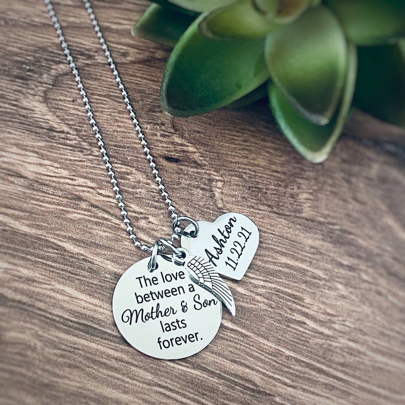 Puede incluir: Collar de plata con dos colgantes. El colgante m&aacute;s grande es redondo y est&aacute; grabado con el texto "The love between a Mother & Son lasts forever." El colgante m&aacute;s peque&ntilde;o es un coraz&oacute;n con el nombre "Ashton" y la fecha "11.22.21" grabados.