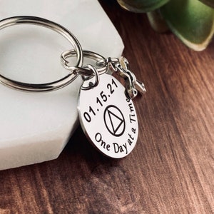 May include: Silver keychain with a round charm featuring the date 01.15.21, a triangle symbol, and the text "One Day at a Time".