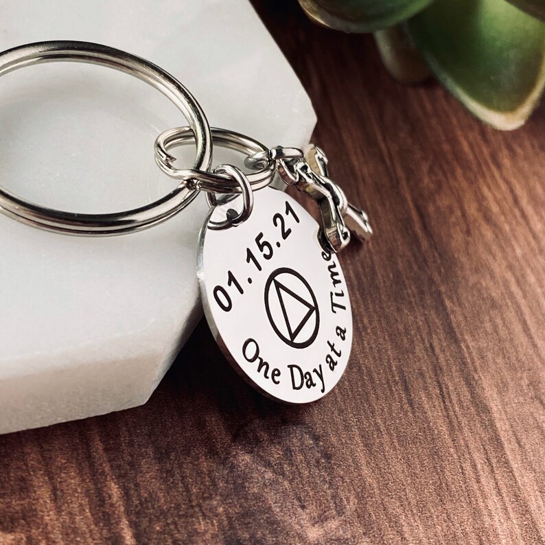 May include: Silver keychain with a round charm featuring the date 01.15.21, a triangle symbol, and the text "One Day at a Time".