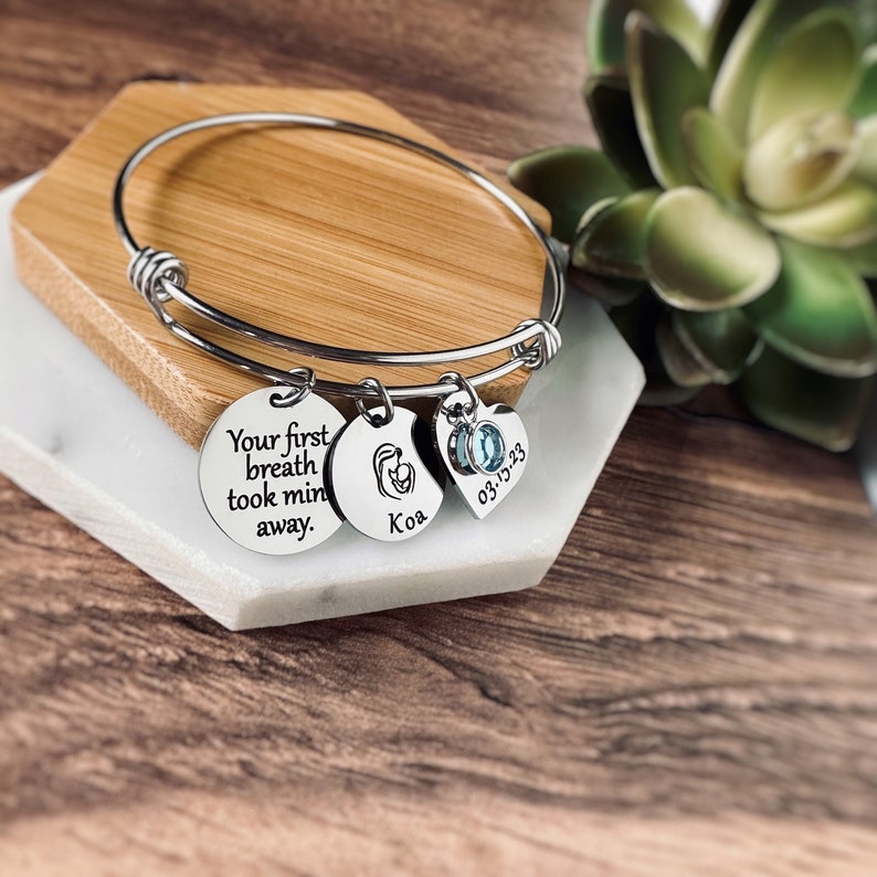 Puede incluir: Un brazalete de plata con tres dijes. Los dijes son un dije redondo con el texto "Your first breath took mine away", un dije redondo con una silueta de beb&eacute; y el texto "Koa", y un dije en forma de coraz&oacute;n con un cristal azul y el texto "03-13-23".