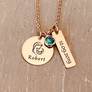 Rose Gold charm necklace with a round disc engraved with an image of a mom holding a baby. Dangling next is a December birthstone. The next charm is a rectangle charm engraved with the date 12.09.19. All charms dangle from a rose gold cable chain.