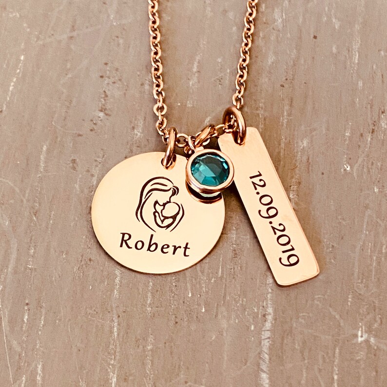 Rose Gold charm necklace with a round disc engraved with an image of a mom holding a baby. Dangling next is a December birthstone. The next charm is a rectangle charm engraved with the date 12.09.19. All charms dangle from a rose gold cable chain.