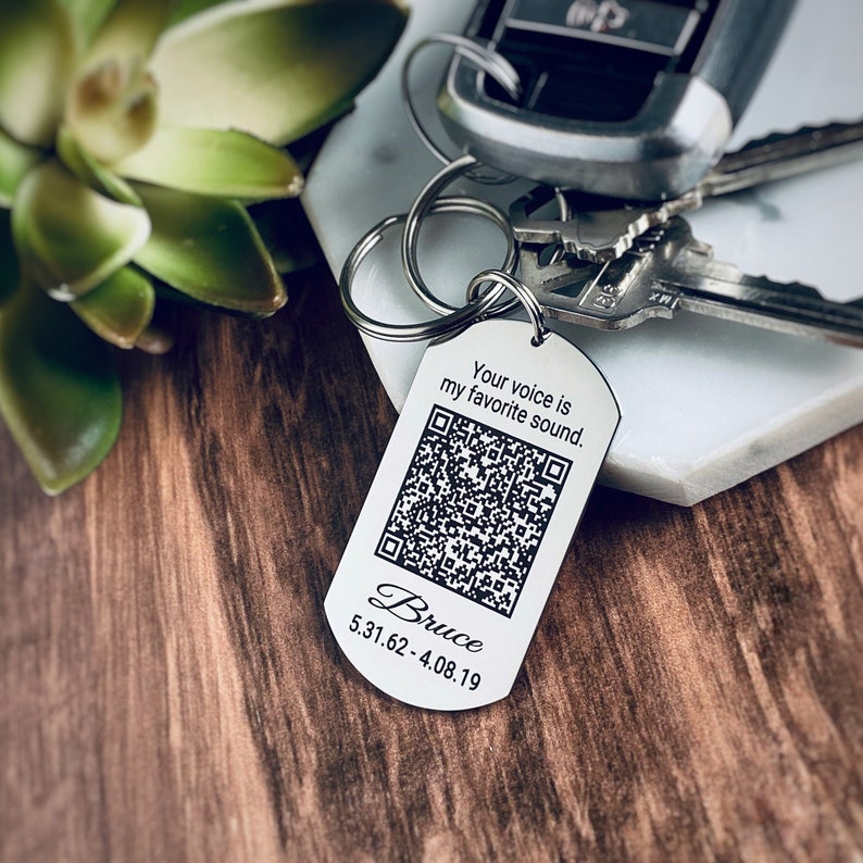 K&ouml;nnte beinhalten: Silberner Schl&uuml;sselanh&auml;nger mit einem schwarz-wei&szlig;en QR-Code, dem Text "Your voice is my favorite sound." und dem Namen "Bruce" mit dem Datum "5.31.62 - 4.08.19".