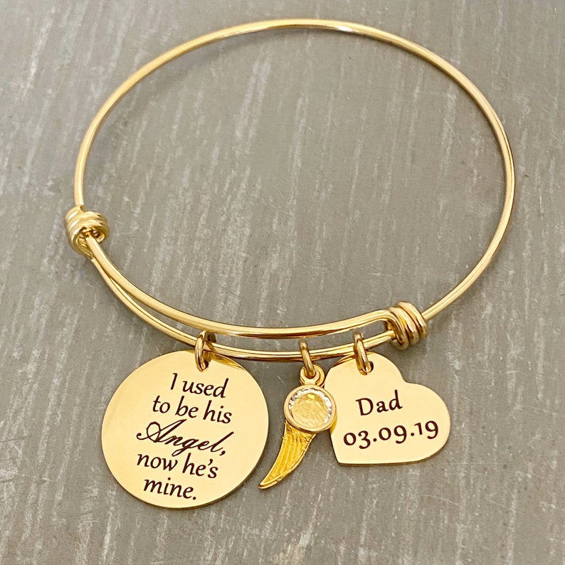 May include: Gold bangle bracelet with three charms. The largest charm is round and engraved with the text "I used to be his Angel, now he's mine". The second charm is a heart-shaped charm with the text "Dad 03.09.19". The third charm is a small wing charm with a clear rhinestone.