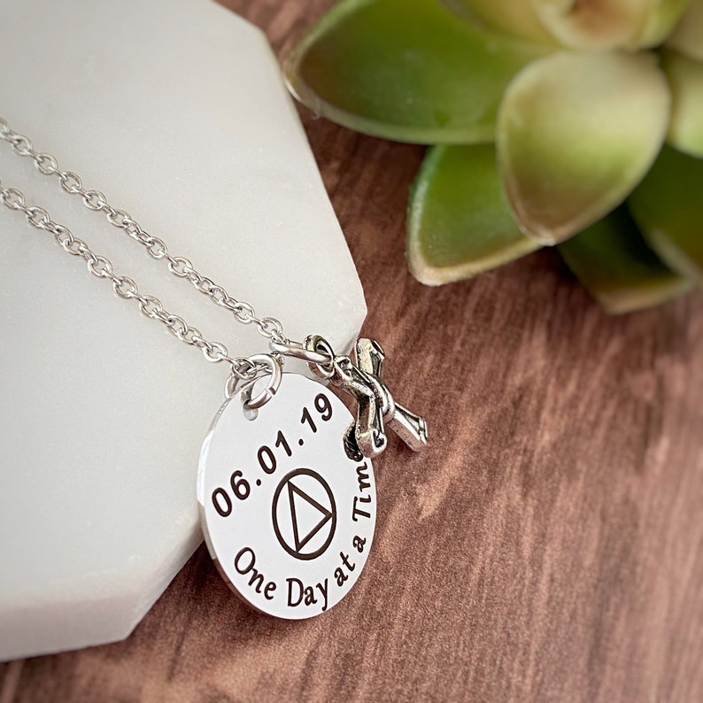 May include: Silver necklace with a round pendant that says "06.01.19 One Day at a Time" with a triangle symbol and a small silver cross charm.