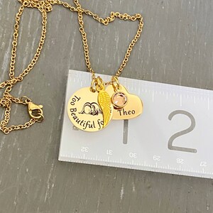 Puede incluir: Collar de oro con tres colgantes: un colgante redondo con el texto "Too Beautiful for Words", un peque&ntilde;o colgante de ala de &aacute;ngel y un colgante de coraz&oacute;n con el nombre "Theo" grabado. El collar se mide con una regla.