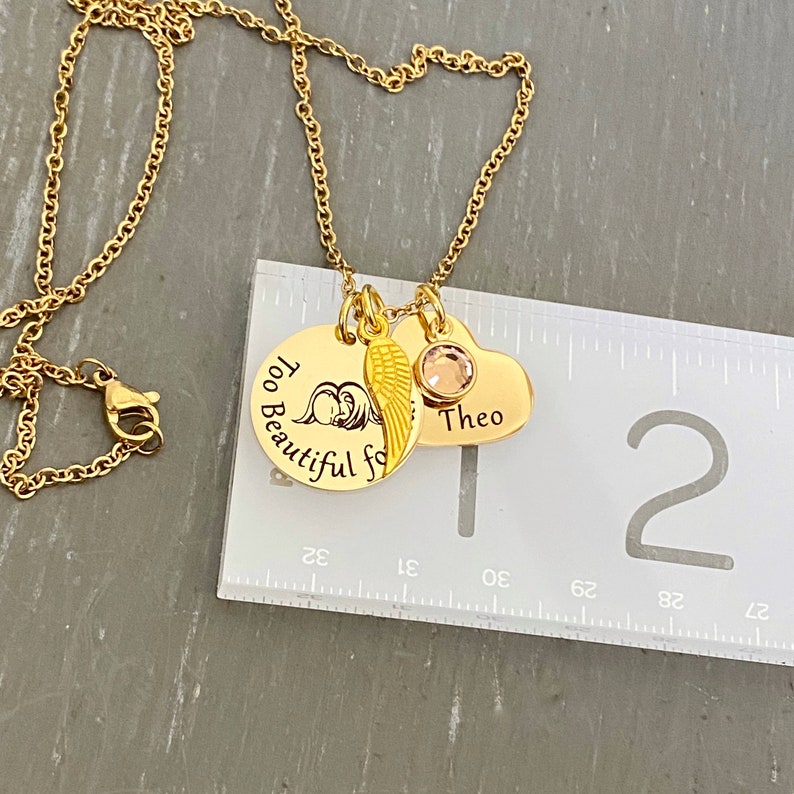 Puede incluir: Collar de oro con tres colgantes: un colgante redondo con el texto "Too Beautiful for Words", un peque&ntilde;o colgante de ala de &aacute;ngel y un colgante de coraz&oacute;n con el nombre "Theo" grabado. El collar se mide con una regla.
