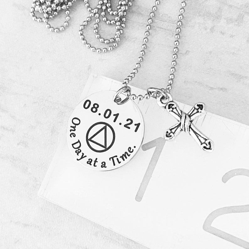 May include: Silver necklace with a round pendant that says "08.01.21 One Day at a Time." The pendant also features a black triangle symbol. The necklace also has a small silver cross charm.