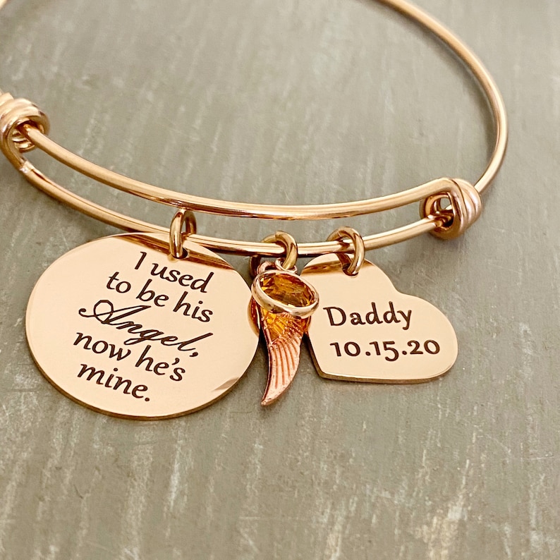 May include: Rose gold bangle bracelet with a round charm that reads "I used to be his Angel, now he's mine." and a heart charm that reads "Daddy 10.15.20". The bracelet also has a small angel wing charm with a clear stone.