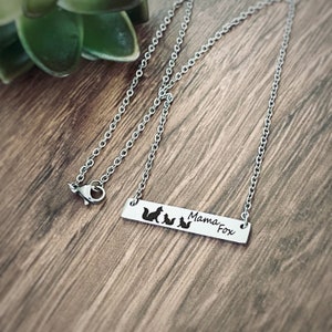 May include: A silver necklace with a silver bar pendant. The pendant has a silhouette of a mama fox and three baby foxes with the text "Mama Fox" on the bar.