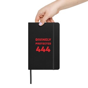 May include: A black hardcover notebook featuring a vertical elastic closure band and a ribbon bookmark. The front cover displays the text DIVINELY PROTECTED and the number 444 printed in a bold, red, sans-serif font centered on the surface.