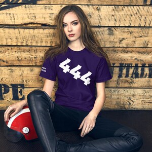 Könnte beinhalten: Ein dunkelviolettes Kurzarm-T-Shirt mit der weißen Zahl 444, die diagonal auf der Brust aufgedruckt ist, und dem Text DIVINELY PROTECTED in Weiß auf dem rechten Ärmel, kombiniert mit einer schwarzen Hose und einem rot-weißen Helm.
