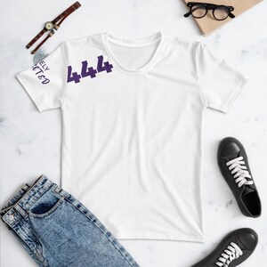 Könnte beinhalten: Ein weißes kurzärmeliges T-Shirt mit der violetten Zahl 444 auf der Brust und dem Text "DIVINELY PROTECTED" in violetter Schrift auf dem linken Ärmel. Das T-Shirt hat einen Rundhalsausschnitt und eine klassische Passform.