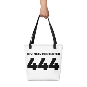 Könnte beinhalten: Eine weiße Tragetasche mit schwarzen Henkeln, die auf der Vorderseite den schwarzen Schriftzug DIVINELY PROTECTED und die große schwarze Zahl 444 aufweist. Die Tasche hat eine rechteckige Form und besteht aus einem Stoff mit schlichtem Design.