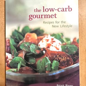 May include: A cookbook titled "the low-carb gourmet: Recipes for the New Lifestyle" by Brigit Binns. The cover features a close-up of a salad with figs, greens, and a creamy dressing.
