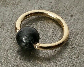 May include: A gold ring with a black gemstone bead. The ring is on a grey surface.