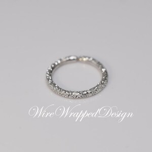 May include: A silver band ring with a row of small, clear stones set around the entire band. The ring is on a white background. "Wire Wrapped Design" is written in cursive below the ring.