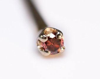 May include: A close-up of a small, red gemstone nose ring. The ring is made of gold and has a simple, elegant design.