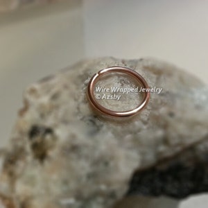 May include: A close-up of a rose gold hoop earring on a white rock. The earring is made of wire and is about 1.2 cm in diameter. The text "Wire Wrapped Jewelry &copy; Azsby" is visible in the image.