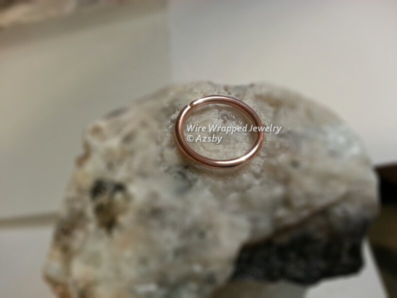 May include: A close-up of a rose gold hoop earring on a white rock. The earring is made of wire and is about 1.2 cm in diameter. The text "Wire Wrapped Jewelry &copy; Azsby" is visible in the image.