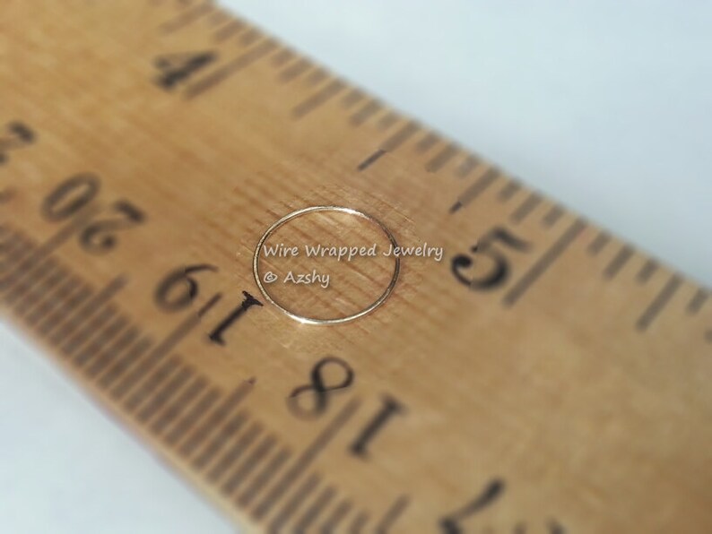 May include: A close-up of a thin gold nose ring, measured against a wooden ruler. The ring is approximately 1/8 inch in diameter. The text "Wire Wrapped Jewelry @Azshy" is visible in the image.