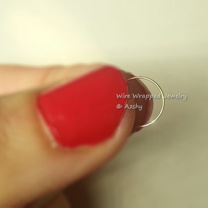 May include: A thin gold hoop nose ring, held between a finger with red nail polish. The ring is a simple, minimalist design.
