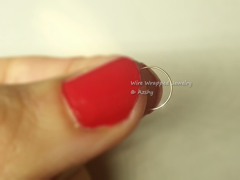 May include: A thin gold hoop nose ring, held between a finger with red nail polish. The ring is a simple, minimalist design.
