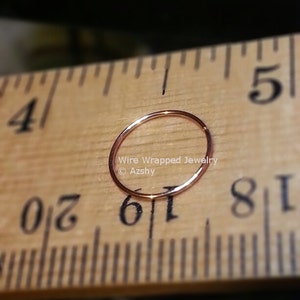 May include: A close-up of a gold-colored nose ring hoop, measured against a wooden ruler. The ring is 1/16 inch in diameter. The text "Wire Wrapped Jewelry &copy; Azshy" is visible in the image.