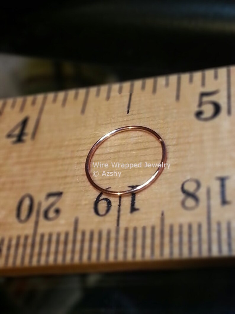 May include: A close-up of a gold-colored nose ring hoop, measured against a wooden ruler. The ring is 1/16 inch in diameter. The text "Wire Wrapped Jewelry &copy; Azshy" is visible in the image.
