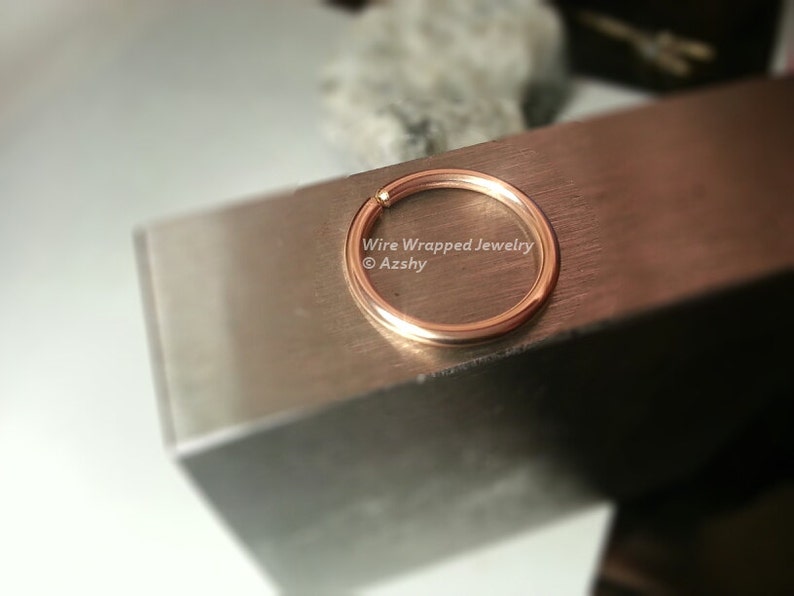 May include: A close-up of a gold-coloured metal ring, likely a nose ring, on a silver surface. The text "Wire Wrapped Jewelry &copy; Azshy" is visible below the ring.