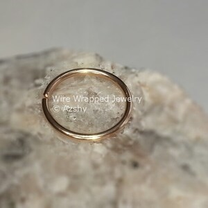 May include: A gold-colored nose ring hoop on a white rock background. The text "Wire Wrapped Jewelry &copy; Azzshy" is visible below the ring.