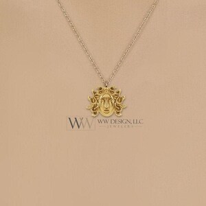 May include: A gold-toned necklace featuring a pendant shaped like a face with snake-like hair, suspended from a delicate chain. The text WW WW DESIGN, LLC. JEWELERS is printed below the pendant in a dark font against the solid background.