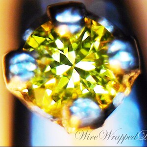May include: A close-up of a green gemstone set in a silver prong setting. The gemstone is faceted and has a bright, sparkling appearance.