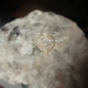 May include: A thin gold wire ring, resting on a white rock. The ring is simple and delicate, with a smooth finish. The text "Wire Wrapped Jewelry &copy; Azshky" is visible below the ring.