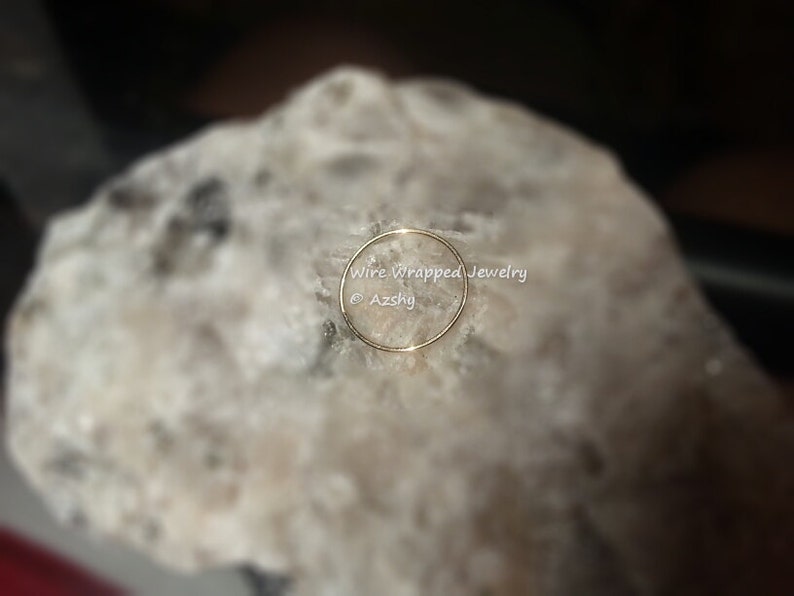 May include: A thin gold wire ring, resting on a white rock. The ring is simple and delicate, with a smooth finish. The text "Wire Wrapped Jewelry &copy; Azshky" is visible below the ring.