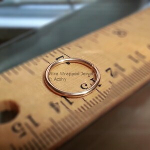 May include: A close-up of a gold-colored nose ring hoop, measured against a wooden ruler. The ring is approximately 1/4 inch in diameter.