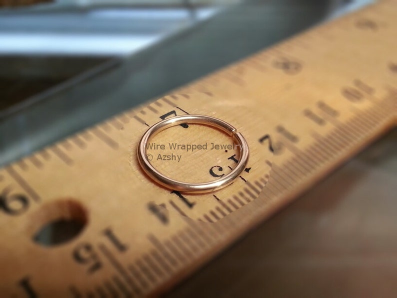 May include: A close-up of a gold-colored nose ring hoop, measured against a wooden ruler. The ring is approximately 1/4 inch in diameter.