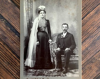 May include: A vintage black and white cabinet card photograph featuring a standing person in a long dark dress with a veil and a seated person in a dark suit. The bottom border includes the text Hutchens Superior Finish Wonewoc Wis.