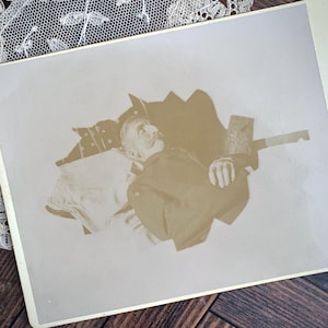 May include: Sepia-toned photograph of a man with a large mustache, lying down. He is wearing a dark jacket and a hat with a decorative pattern. The image is framed with a white border and a lace trim.
