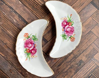May include: Two white porcelain crescent-shaped dishes featuring a floral pattern with pink roses, orange blossoms, and green leaves. Each dish has a scalloped edge and a thin gold trim along the rim, creating a decorative set of curved plates.